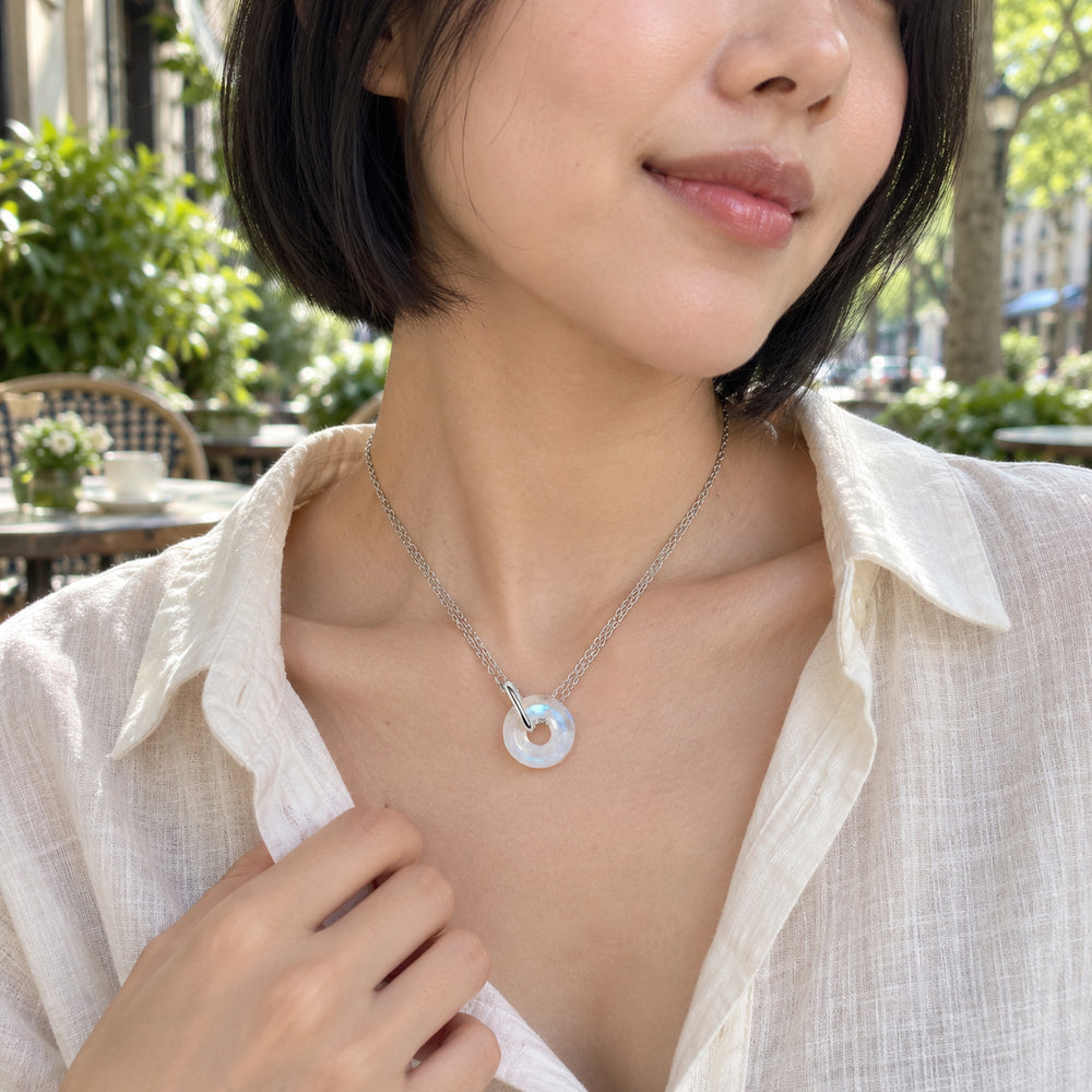 A woman in a cream linen shirt wears a silver double-chain necklace with a round moonstone pendant at an outdoor cafe.