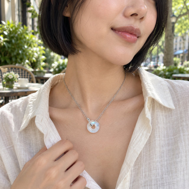 A woman in a cream linen shirt wears a silver double-chain necklace with a round moonstone pendant at an outdoor cafe.