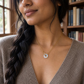 A small open-centre moonstone pendant hangs from two yellow gold vermeil chains above a taupe knit neckline beside a bookcase.