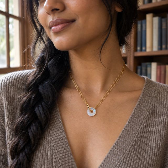 A small open-centre moonstone pendant hangs from two yellow gold vermeil chains above a taupe knit neckline beside a bookcase.