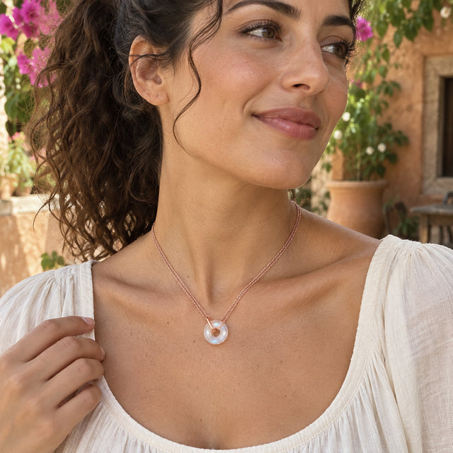 A rose gold vermeil double-chain necklace with a circular moonstone pendant rests above an ivory blouse in a flower-filled courtyard.