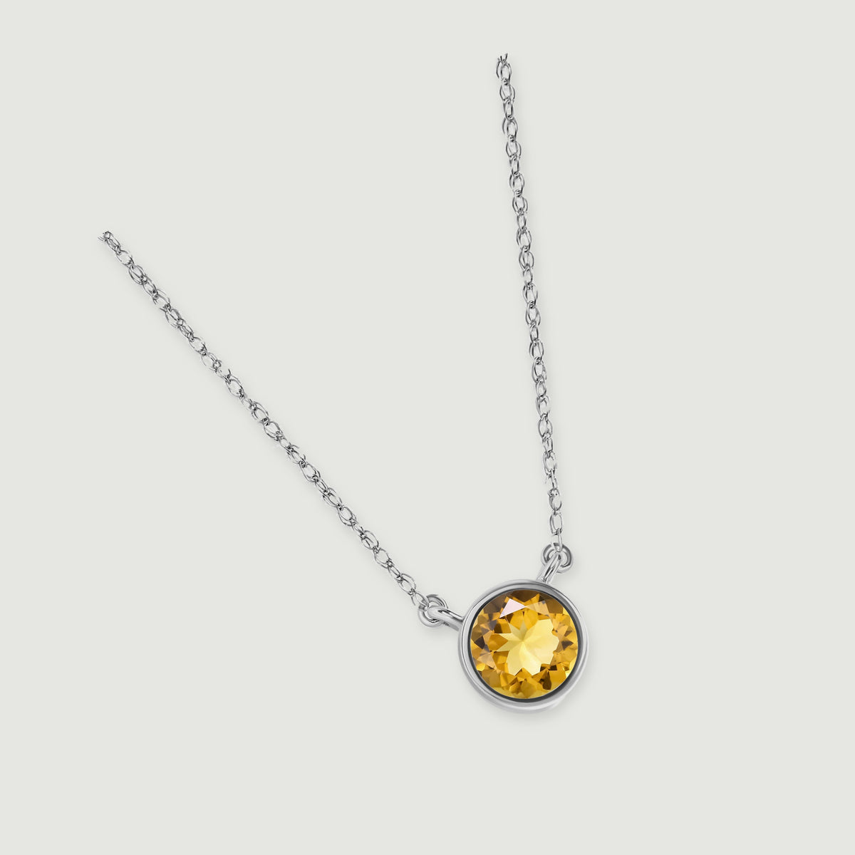 The white gold necklace lies at an angle on a light grey surface with its yellow citrine pendant near the lower right.