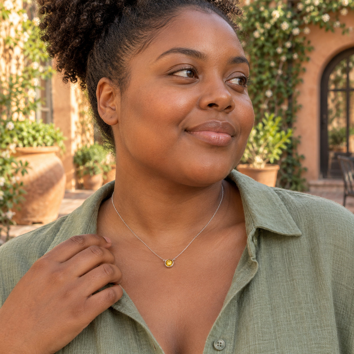 A woman with curly hair wears the yellow citrine pendant on a fine white gold chain above a sage linen shirt in a leafy courtyard.