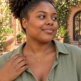 A woman with curly hair wears the yellow citrine pendant on a fine white gold chain above a sage linen shirt in a leafy courtyard.