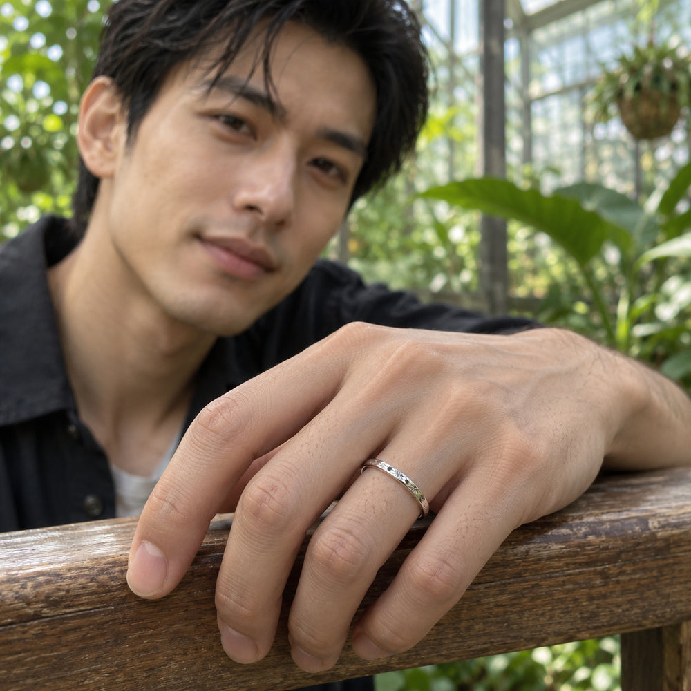 An engraved silver band is worn on a hand resting on a wooden rail in a leafy greenhouse.