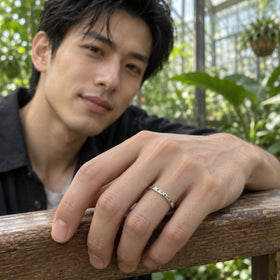 An engraved silver band is worn on a hand resting on a wooden rail in a leafy greenhouse.