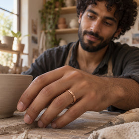 A rose gold band is shown on a hand beside an unglazed clay bowl in a ceramics studio.
