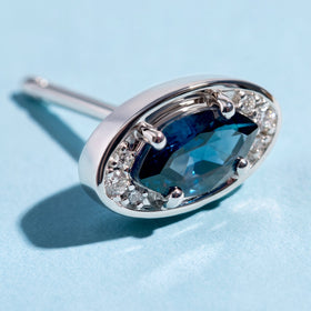 A close view of the silver-tone earring reveals four rounded prongs and clear stones beside a faceted blue center.