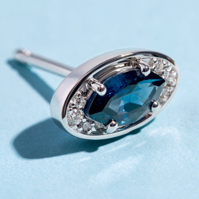 A close view of the silver-tone earring reveals four rounded prongs and clear stones beside a faceted blue center.
