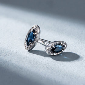 Two silver-tone blue-stone studs rest at different angles on a pale textured surface.