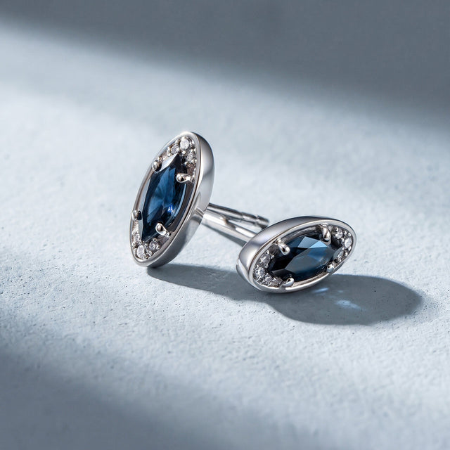 Two silver-tone blue-stone studs rest at different angles on a pale textured surface.