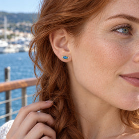 A red-haired woman with freckles wears a yellow-gold-tone blue stud beside a sunlit harbor.