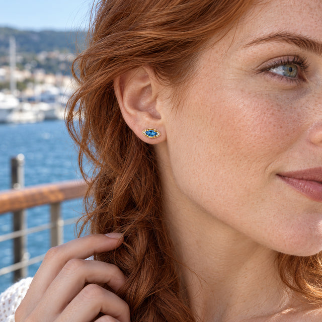 A red-haired woman with freckles wears a yellow-gold-tone blue stud beside a sunlit harbor.