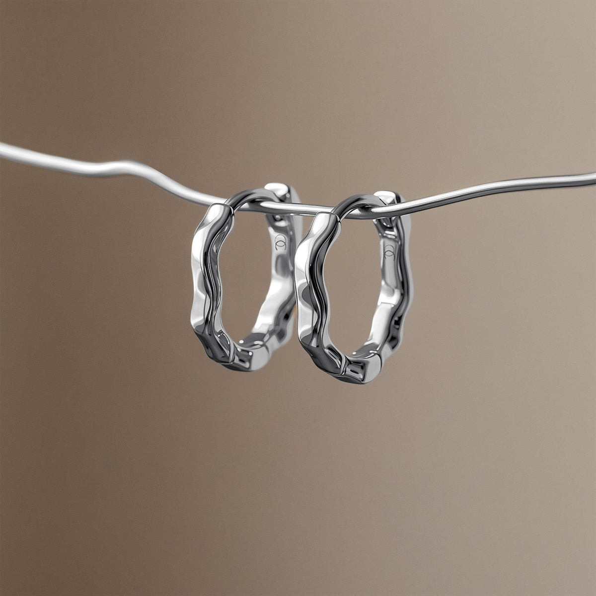 Two sculpted hoop earrings hang from a slender wavy line against a taupe background.