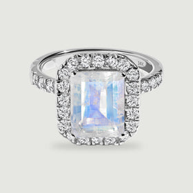 A white gold ring displays a blue-flashing rectangular moonstone within a border of round diamonds.