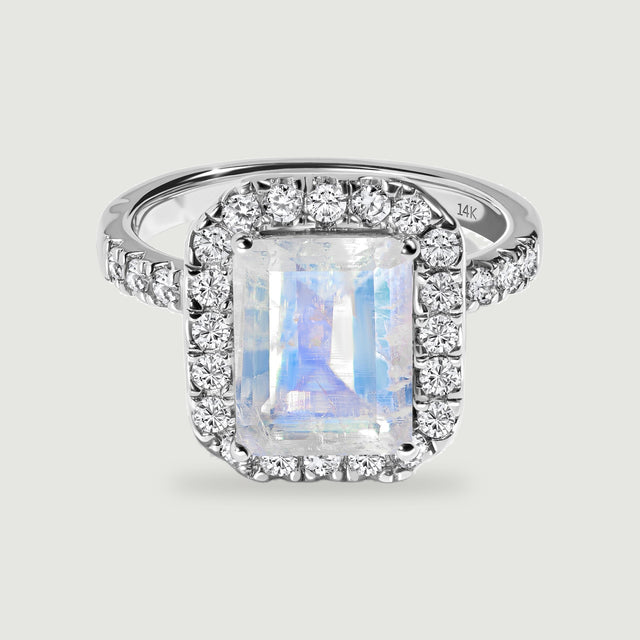 A white gold ring displays a blue-flashing rectangular moonstone within a border of round diamonds.