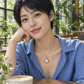 A woman in a blue open-neck shirt wears the white rhodium plated mother of pearl necklace beside cafe windows and greenery.