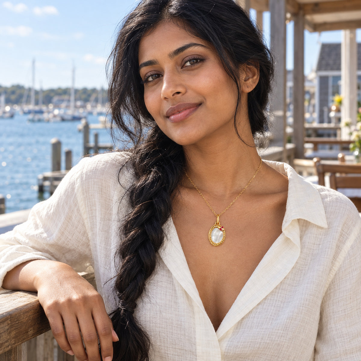 A woman with a dark braid wears the yellow gold plated mother of pearl necklace with an ivory blouse by a harbor railing.