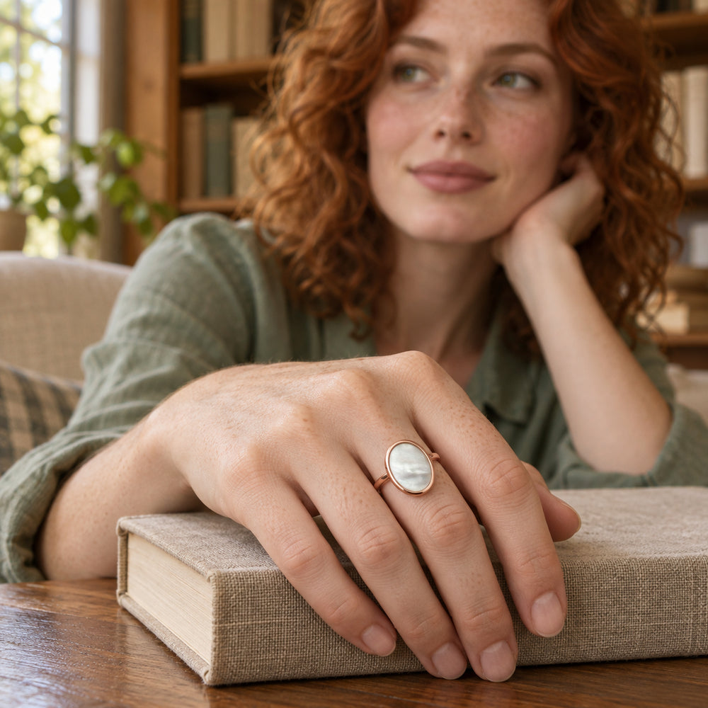 An oval mother-of-pearl ring in rose gold sits on a freckled hand resting on a linen-covered book.