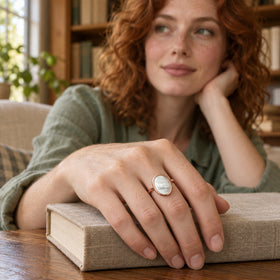 An oval mother-of-pearl ring in rose gold sits on a freckled hand resting on a linen-covered book.