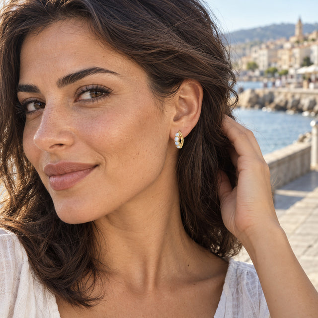 A woman with wavy brown hair wears a yellow-gold-tone moonstone hoop beside the sea.