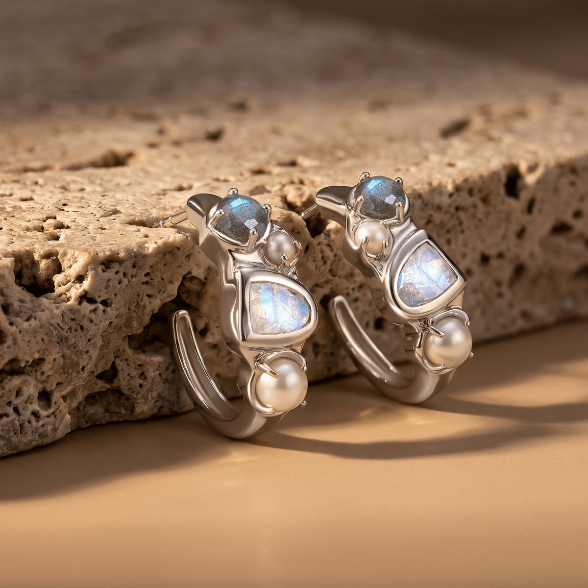 Silver-tone pearl and stone hoop earrings rest upright beside a textured beige stone slab.