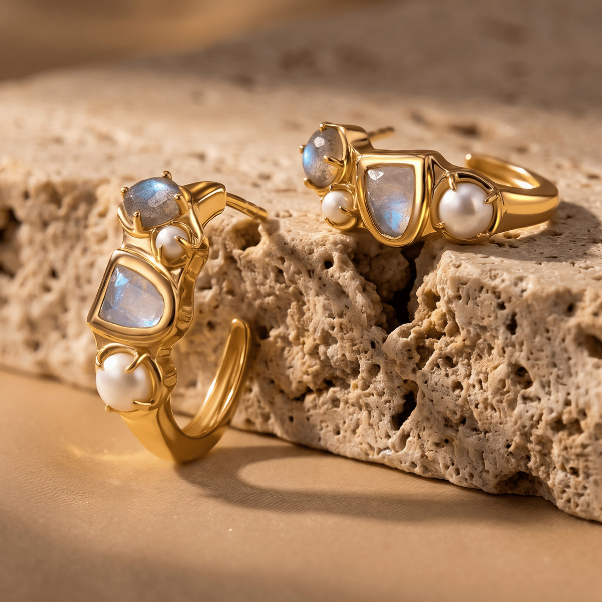 A pair of yellow-gold-tone hoops lies at different angles on a beige stone surface, revealing the curved bands and pearl settings.