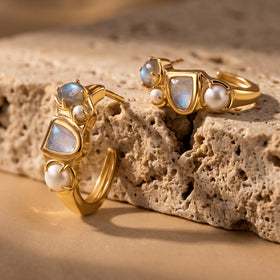 A pair of yellow-gold-tone hoops lies at different angles on a beige stone surface, revealing the curved bands and pearl settings.