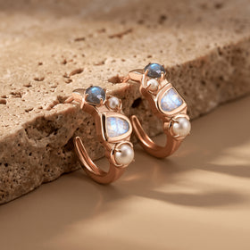 Rose-gold-tone earrings lean against a pale textured stone block with their open hoops and white pearls visible.