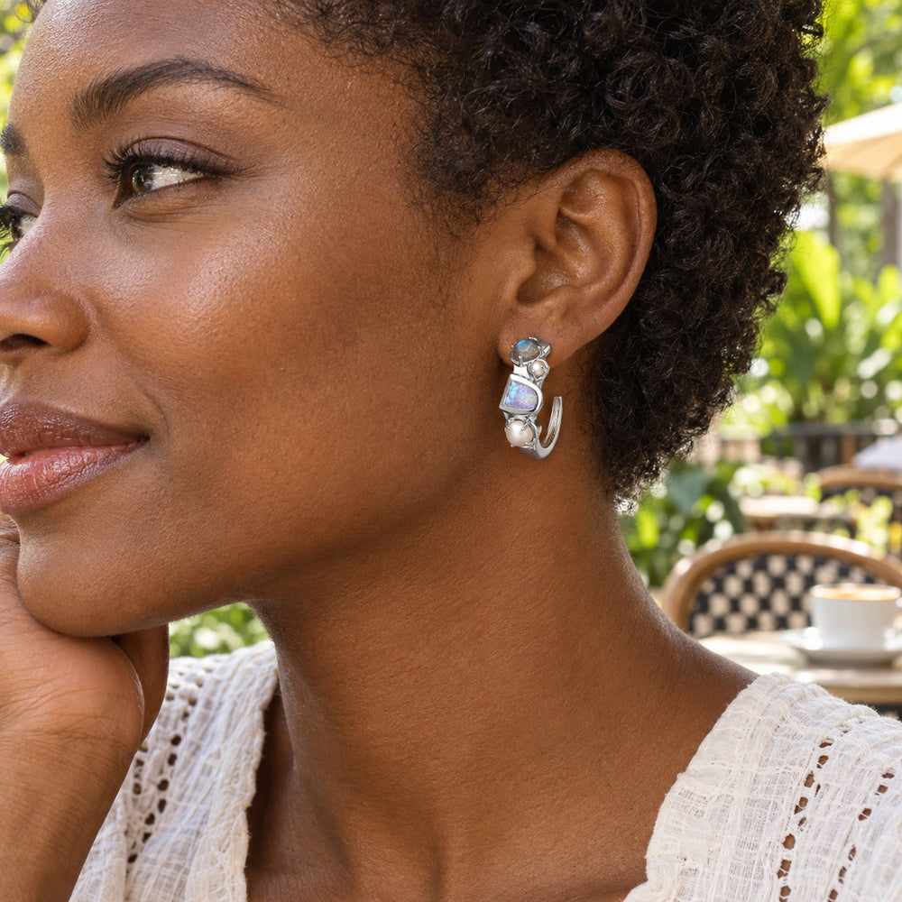 A woman with short natural curls wears a silver-tone pearl and stone hoop earring at a leafy outdoor cafe.