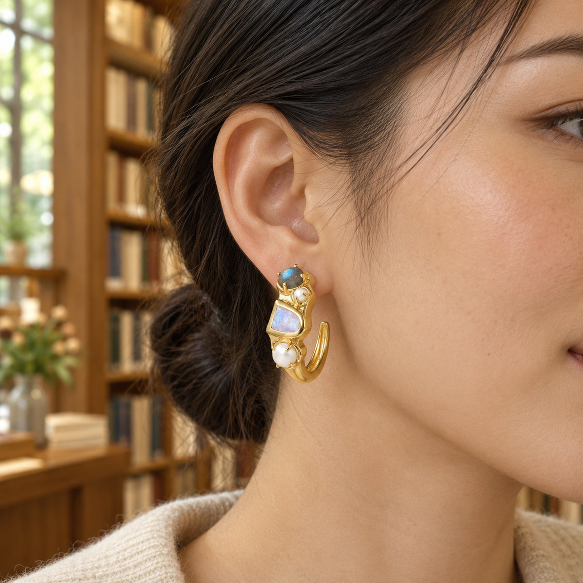 A yellow-gold-tone hoop with white pearls and blue-flashing stones is shown on an ear beside wooden bookshelves.