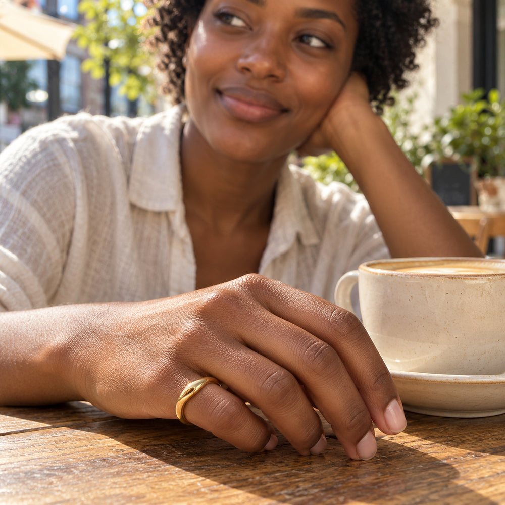 A woman wears the curved yellow gold vermeil ring on her little finger beside a ceramic coffee cup at a wooden cafe table.