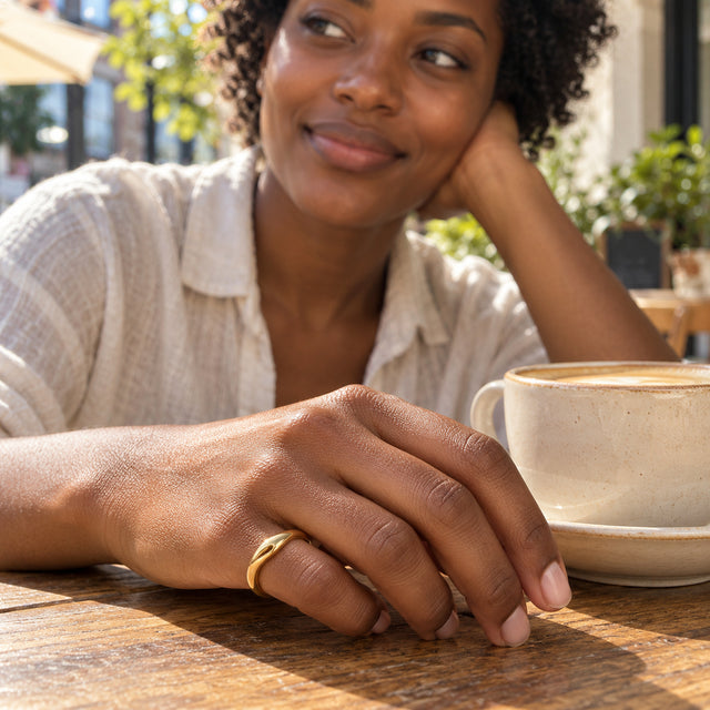 A woman wears the curved yellow gold vermeil ring on her little finger beside a ceramic coffee cup at a wooden cafe table.