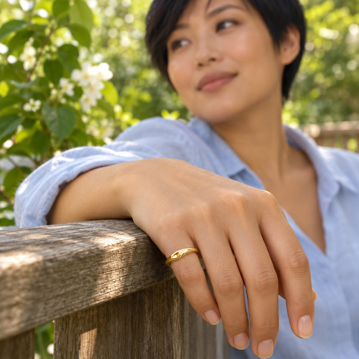 A woman in a pale blue shirt wears the yellow gold vermeil ring on her little finger while resting her hand on a wooden garden railing.