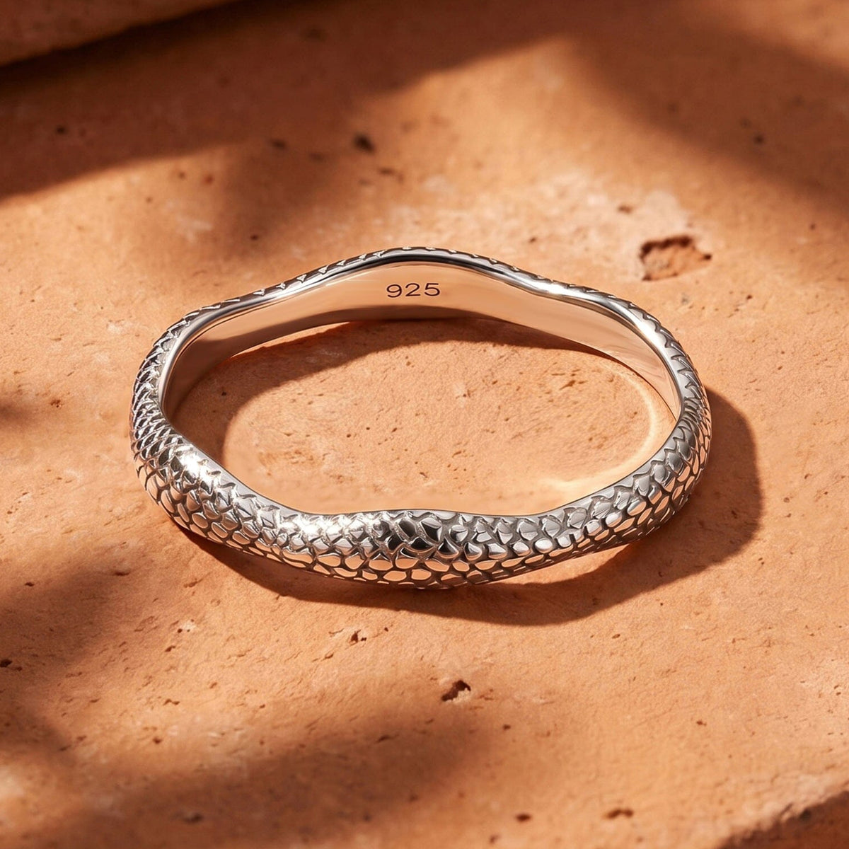 A silver ring lies on a warm stone surface with sunlight crossing its raised texture.