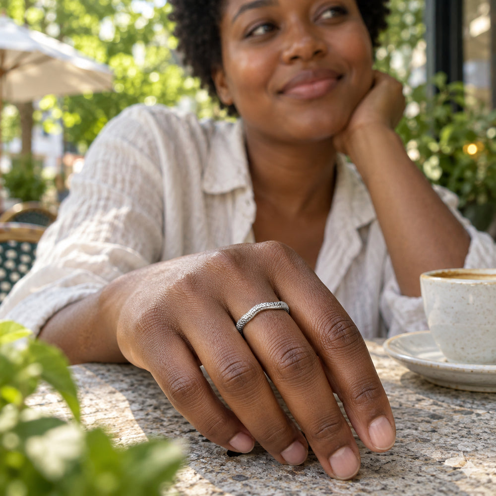 A textured silver band is worn on a hand resting on a stone cafe table beside a ceramic cup.