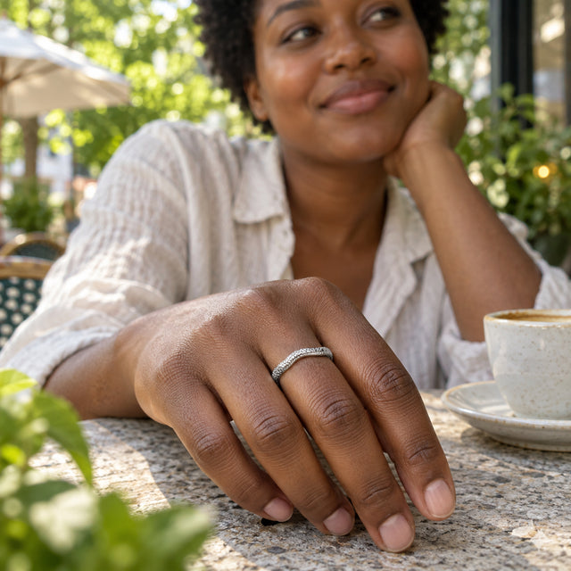 A textured silver band is worn on a hand resting on a stone cafe table beside a ceramic cup.