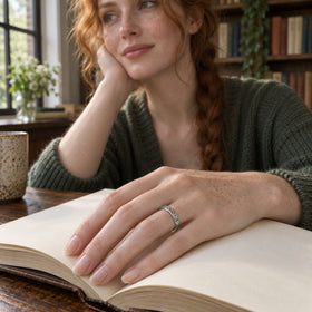 A silver band with a rippled edge is shown on a freckled hand resting across an open book.