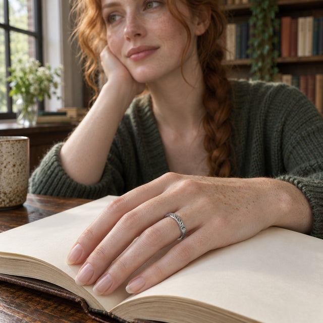A silver band with a rippled edge is shown on a freckled hand resting across an open book.