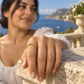 A textured yellow gold band is worn on an index finger above a sunlit stone terrace and blue water.