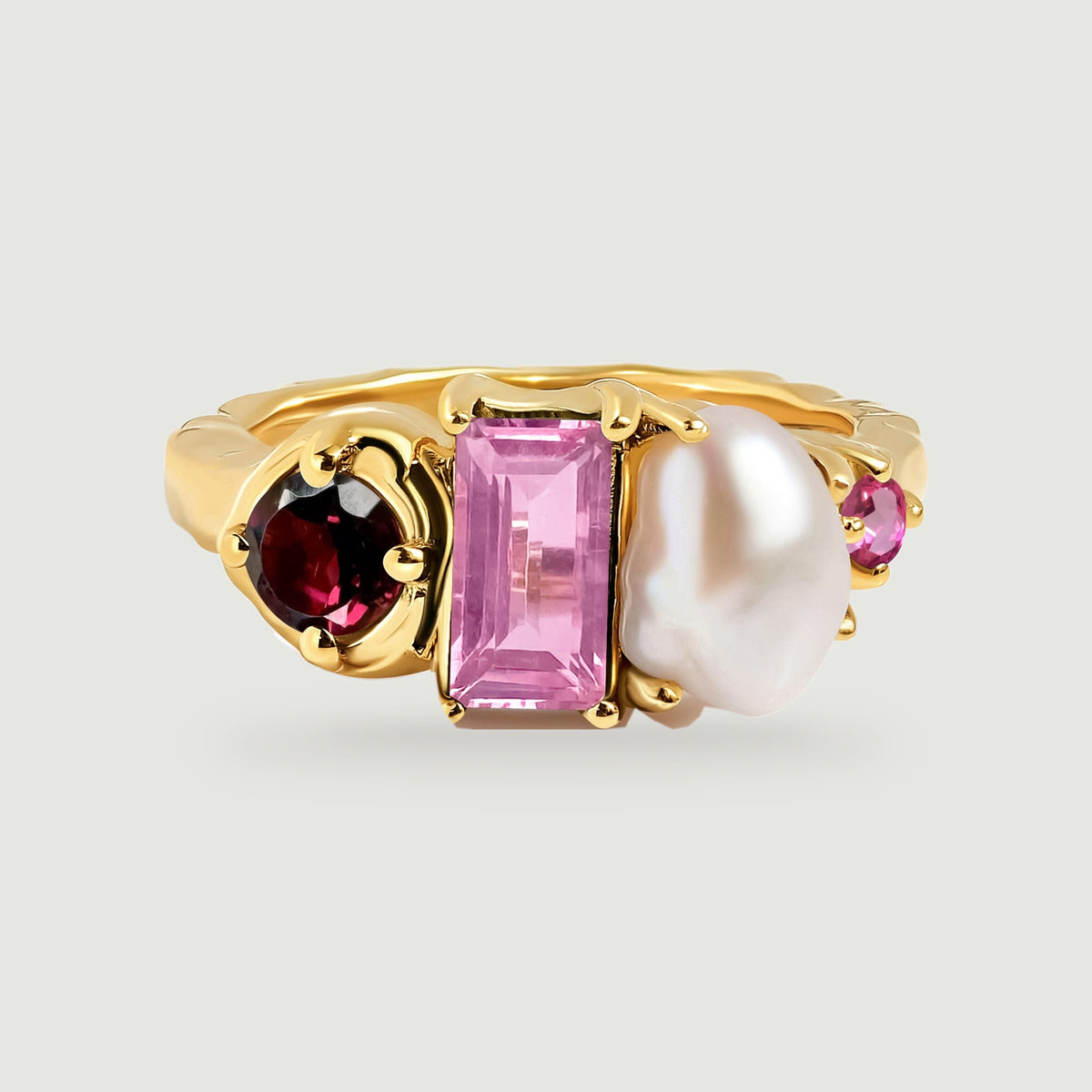 A yellow gold ring displays a deep red round stone, a rectangular pink stone, an irregular white pearl, and a small pink accent on a pale gray background.