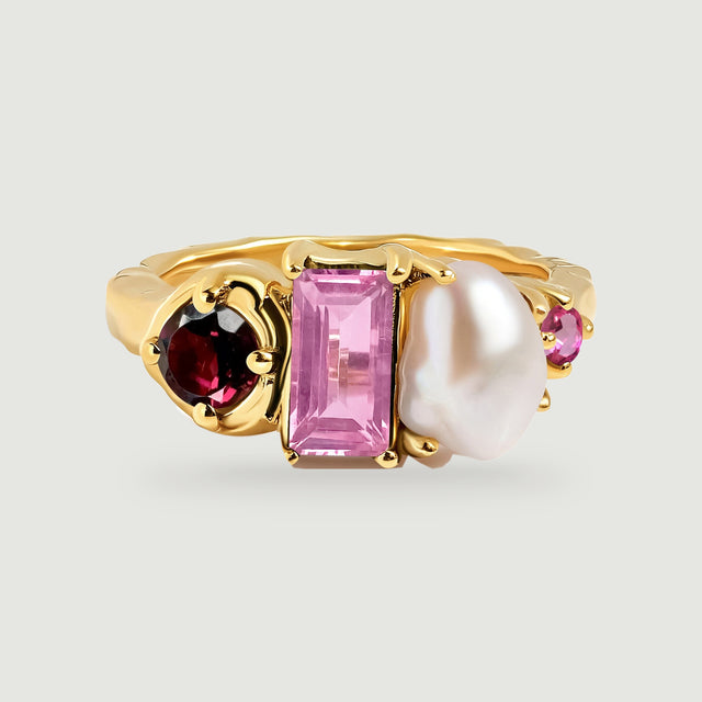 A yellow gold ring displays a deep red round stone, a rectangular pink stone, an irregular white pearl, and a small pink accent on a pale gray background.