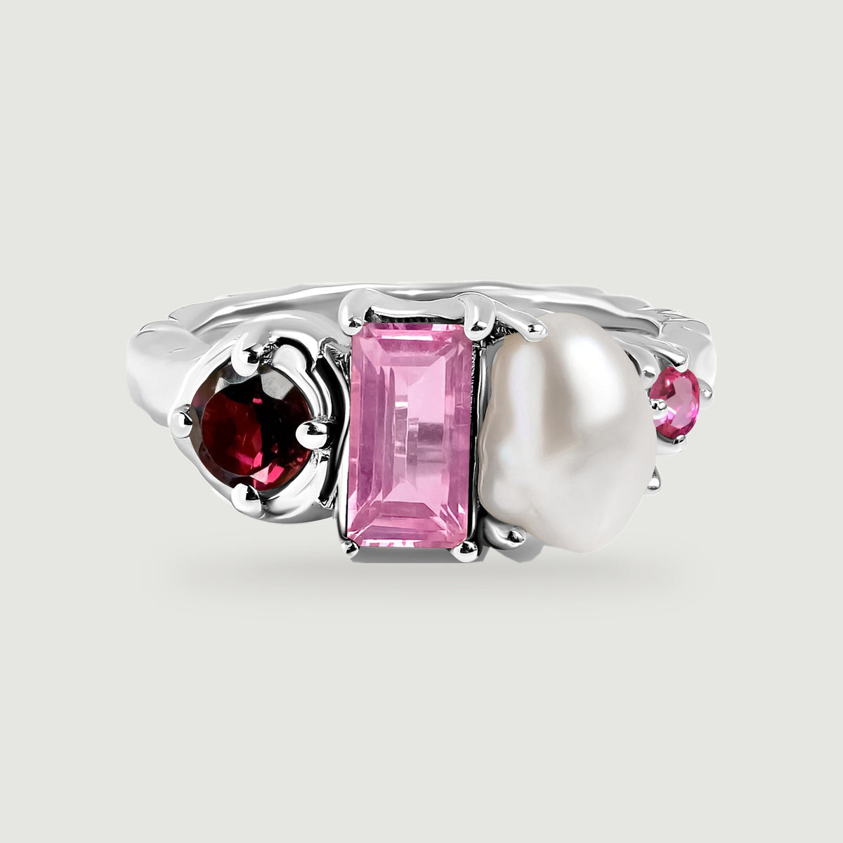 A polished silver ring with rounded prongs surrounds red and pink faceted stones beside a lustrous white pearl.