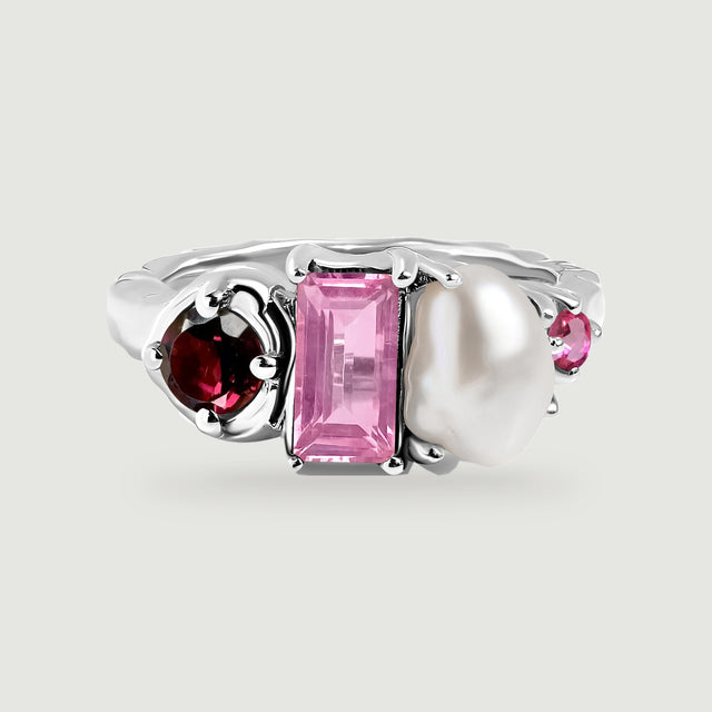 A polished silver ring with rounded prongs surrounds red and pink faceted stones beside a lustrous white pearl.