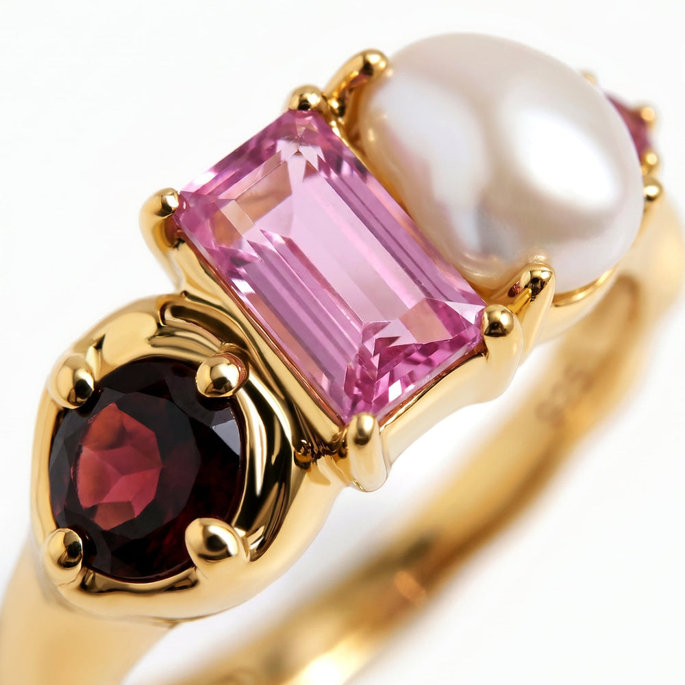 The close view of a yellow gold ring reveals the facets of its dark red round stone, rectangular pink stone, and irregular pearl.