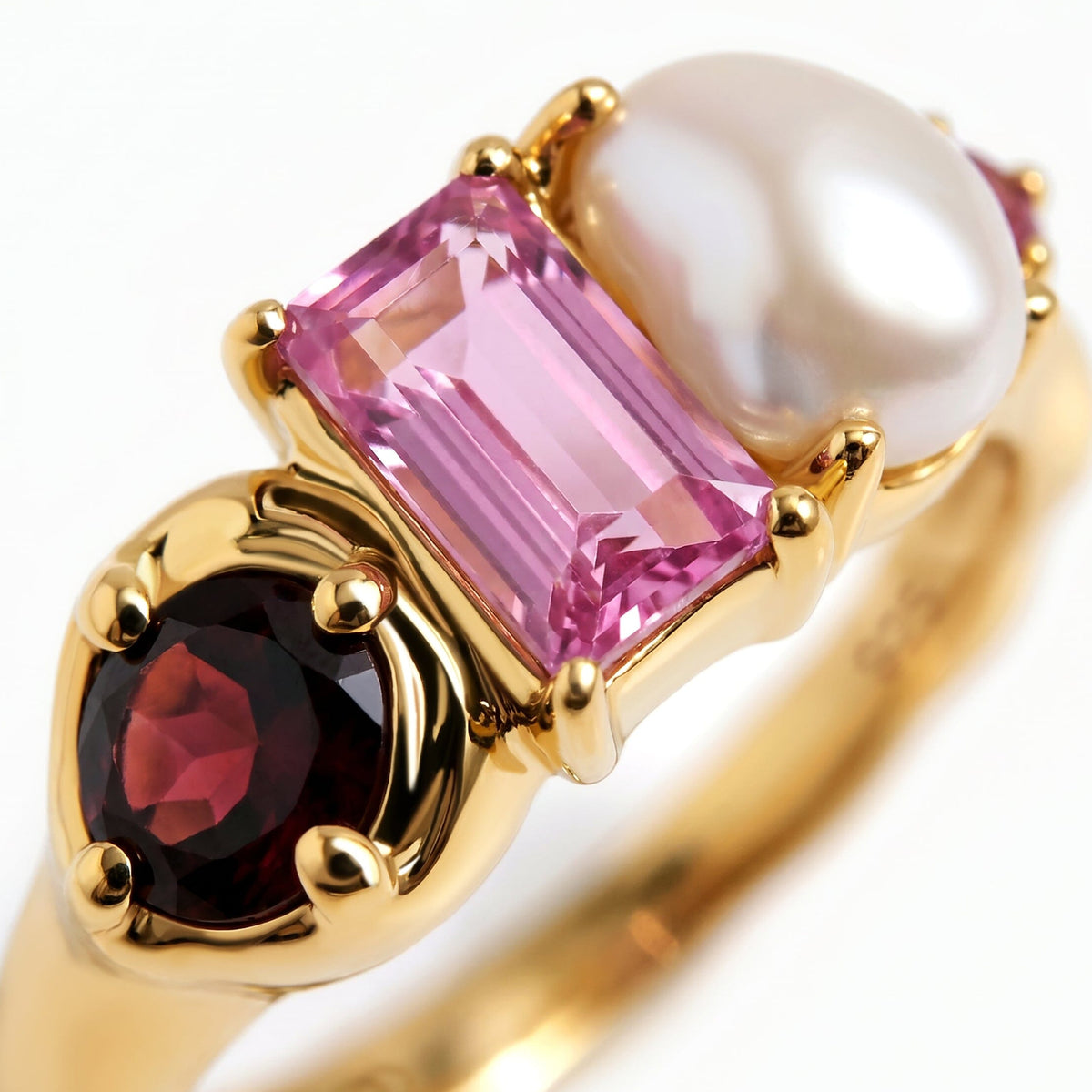 The close view of a yellow gold ring reveals the facets of its dark red round stone, rectangular pink stone, and irregular pearl.
