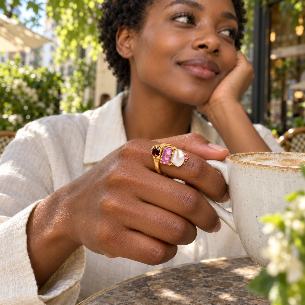 A woman with short curly hair wears a yellow gold gemstone and pearl ring while holding a ceramic coffee cup on a leafy cafe terrace.