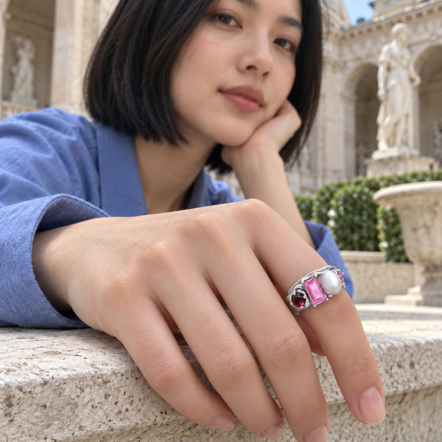 A woman in a blue sleeve shows a silver ring with red and pink stones and a white pearl against a stone courtyard bench.