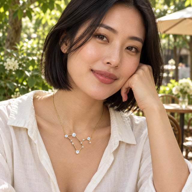 A woman in a cream linen shirt wears the yellow gold moonstone necklace at an outdoor cafe with leafy plants behind her.