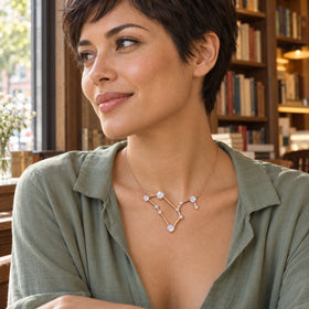 A woman with short dark hair wears the rose gold necklace above a sage blouse beside a window and wooden bookshelves.